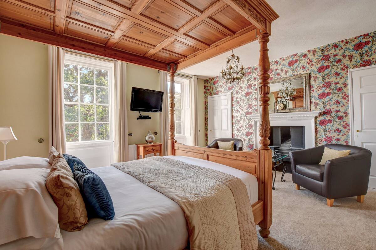 A four-poster bed with a wooden headboard, a television mounted on the wall, a fireplace with a mirror above it, and a floral wallpaper.