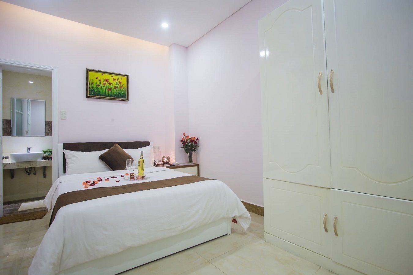 A hotel room with a bed, a nightstand with a lamp, a mirror, a wardrobe, and a painting on the wall.