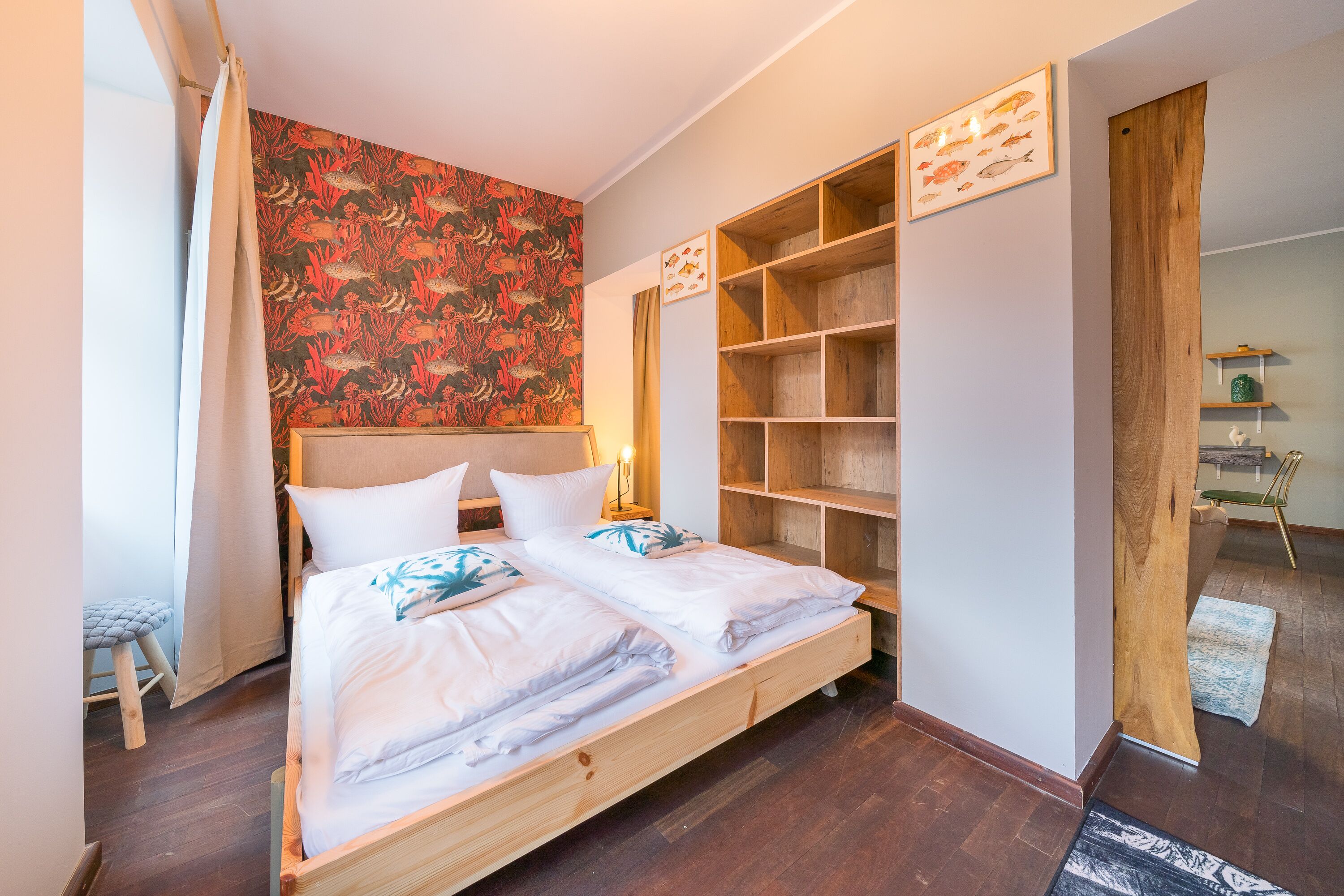 A double bed with white linens, a wooden headboard, and a coral-patterned wall. There are built-in wooden shelves and a small stool.