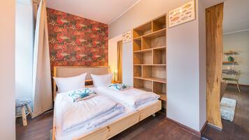 A double bed with white linens, a wooden headboard, and a coral-patterned wall. There are built-in wooden shelves and a small stool.