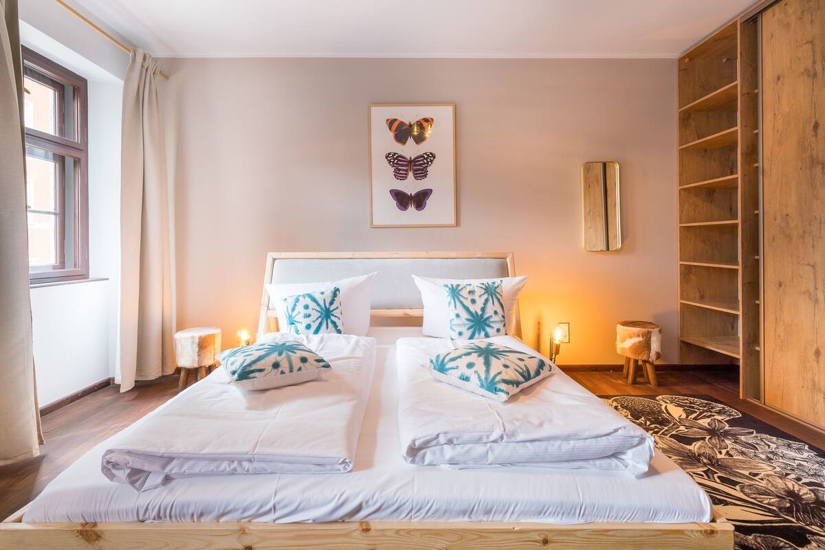 Ein Doppelbett mit weißen Leinen und türkisfarbenen Kissen mit Muster, ein hölzerner Kopfteil, ein gerahmtes Schmetterlingsbild über dem Bett, ein Holzschrank und ein Teppich mit Muster.