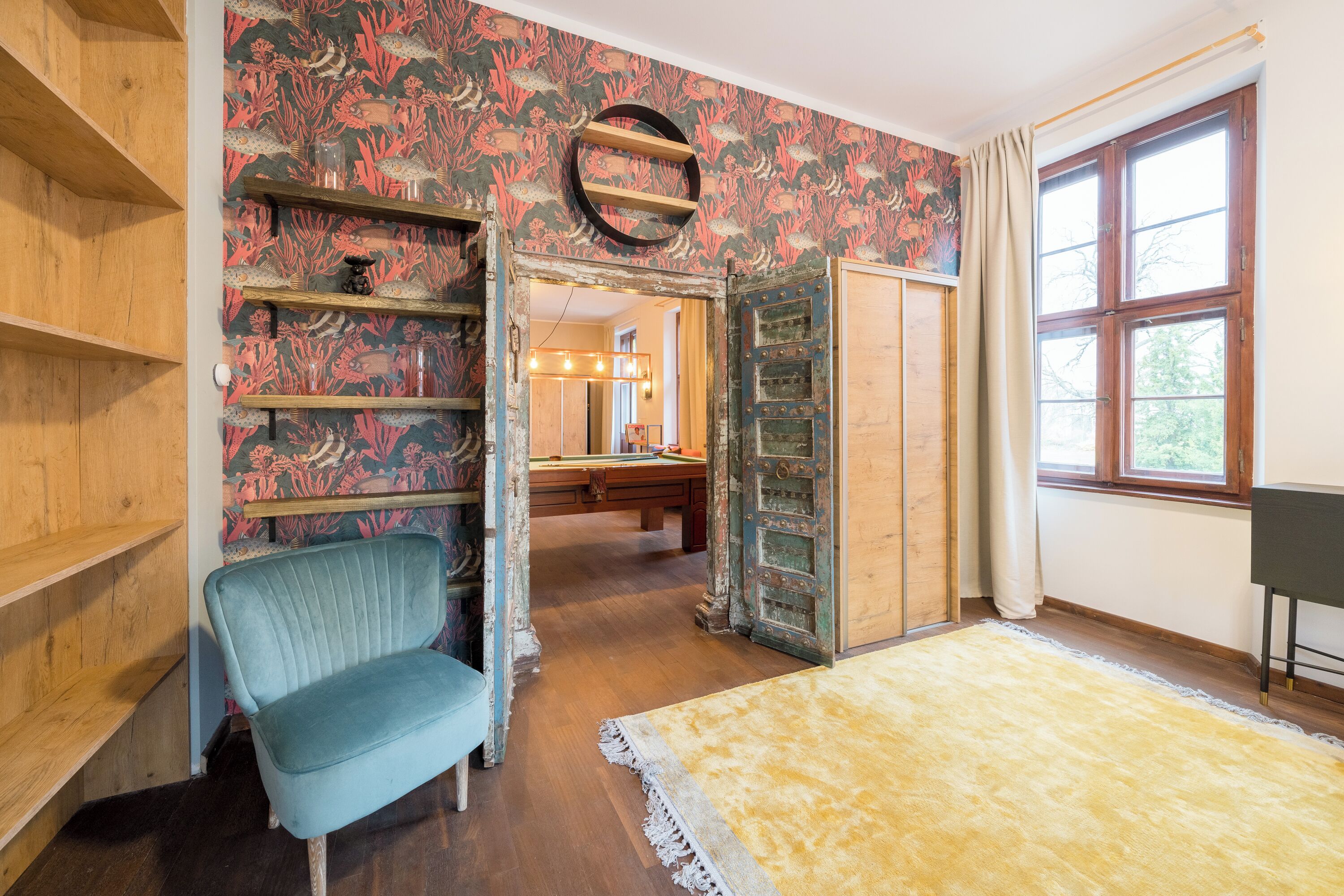 A room with wooden shelves, a blue chair, a patterned wallpaper, a wooden door, and a window with curtains.
