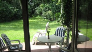 Un'area patio con un tavolo rotondo e due sedie a righe, una piccola pianta in vaso e vista su un prato curato e alberi.