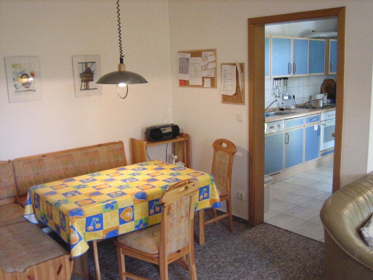 A dining area with a table covered in a checkered tablecloth, wooden chairs, a hanging lamp, and a bulletin board on the wall.