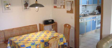 A dining area with a table covered in a checkered tablecloth, wooden chairs, a hanging lamp, and a bulletin board on the wall.