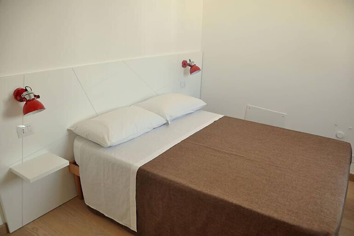 A bedroom with a bed, white walls, and two red wall-mounted lamps.