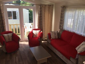 A living room with a red sofa, two red armchairs, a wooden coffee table, and a sliding glass door leading to an outdoor area with a white chair. - La Sensation (Matringhem)