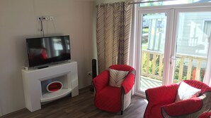 Un salon avec un téléviseur à écran plat posé sur un meuble blanc, deux fauteuils rouges, une porte vitrée donnant sur un balcon et un plancher en bois.