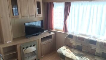 A compact RV interior with a flat-screen TV, wooden cabinets, a small kitchenette, and a window with curtains.