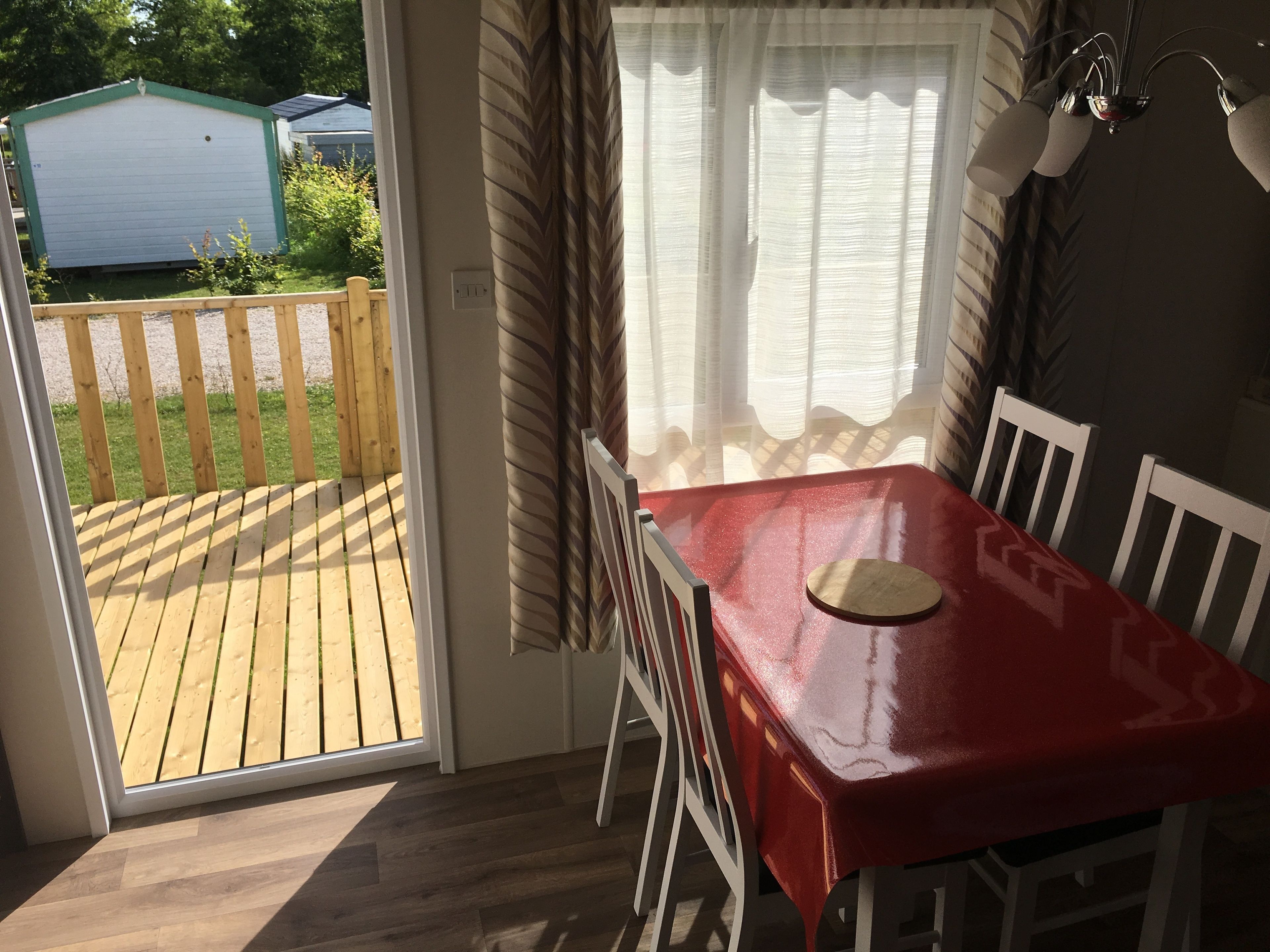 A room with a wooden deck, a red table, white chairs, and a window with sheer curtains.