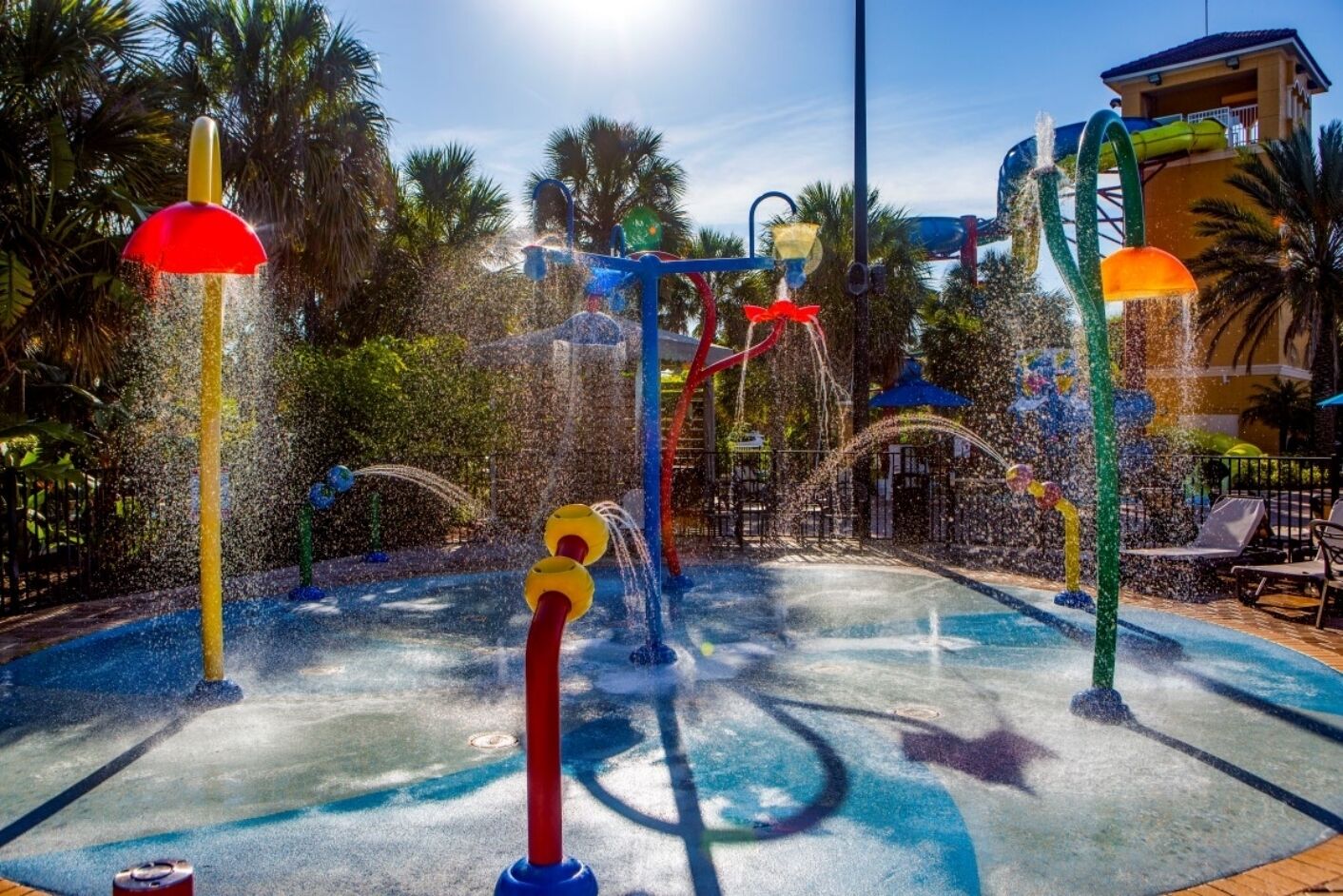 A water play area with multiple water jets and slides, surrounded by palm trees and a pool.