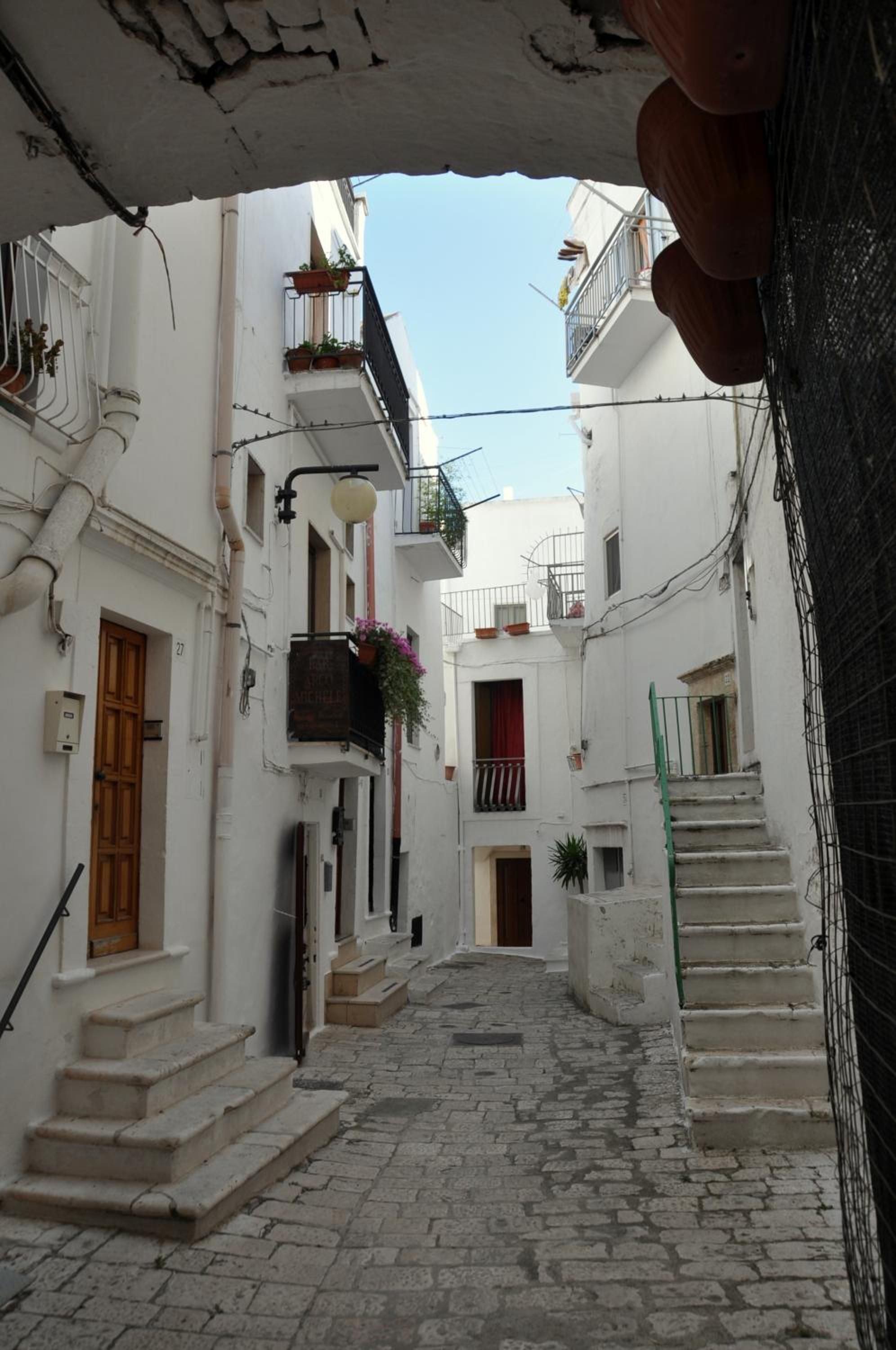 A narrow cobblestone street flanked by white buildings with balconies, a wooden door, and a staircase leading to an upper level.
