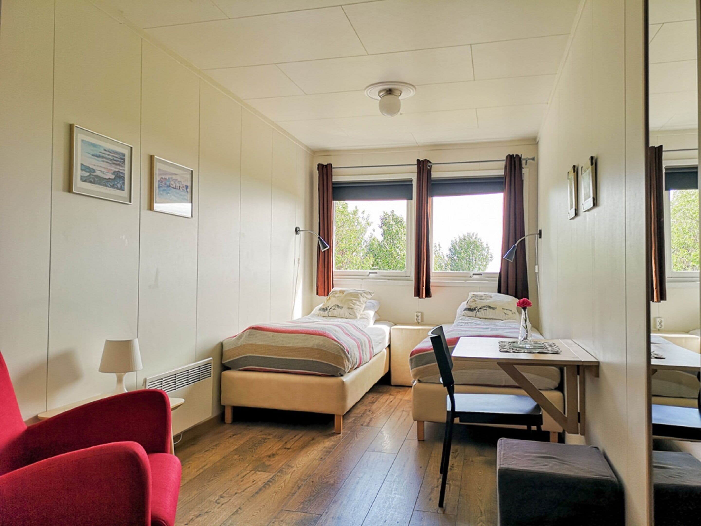 A small, well-lit room with two beds, a desk, a chair, and a red armchair.