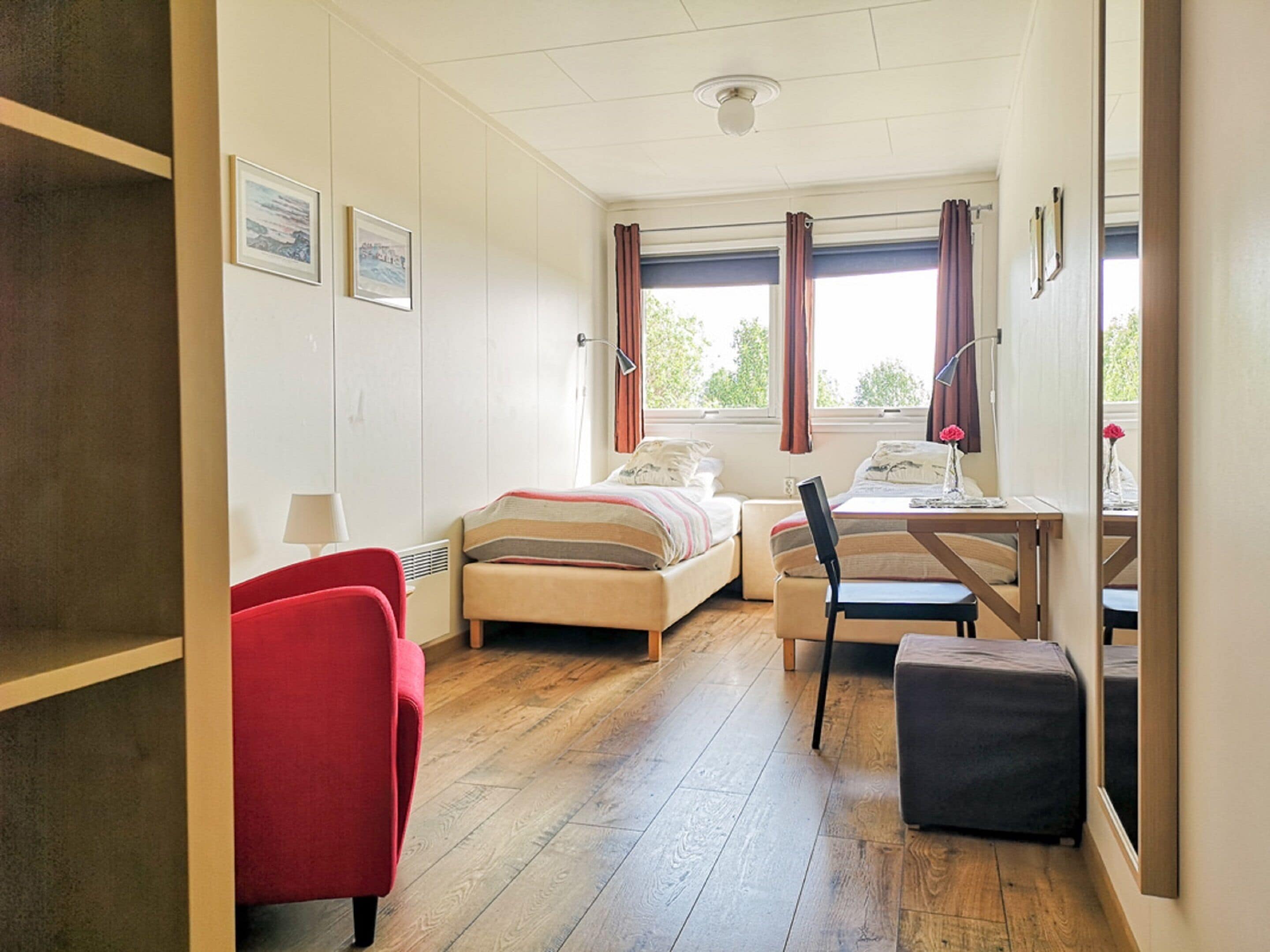 A compact hotel room with a bed, a desk with a chair, a red armchair, and a small table with a lamp.
