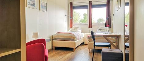 A compact hotel room with a bed, a desk with a chair, a red armchair, and a small table with a lamp.
