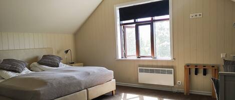 A bedroom with a sloped ceiling, a large window, a bed, a bedside table, and a radiator.
