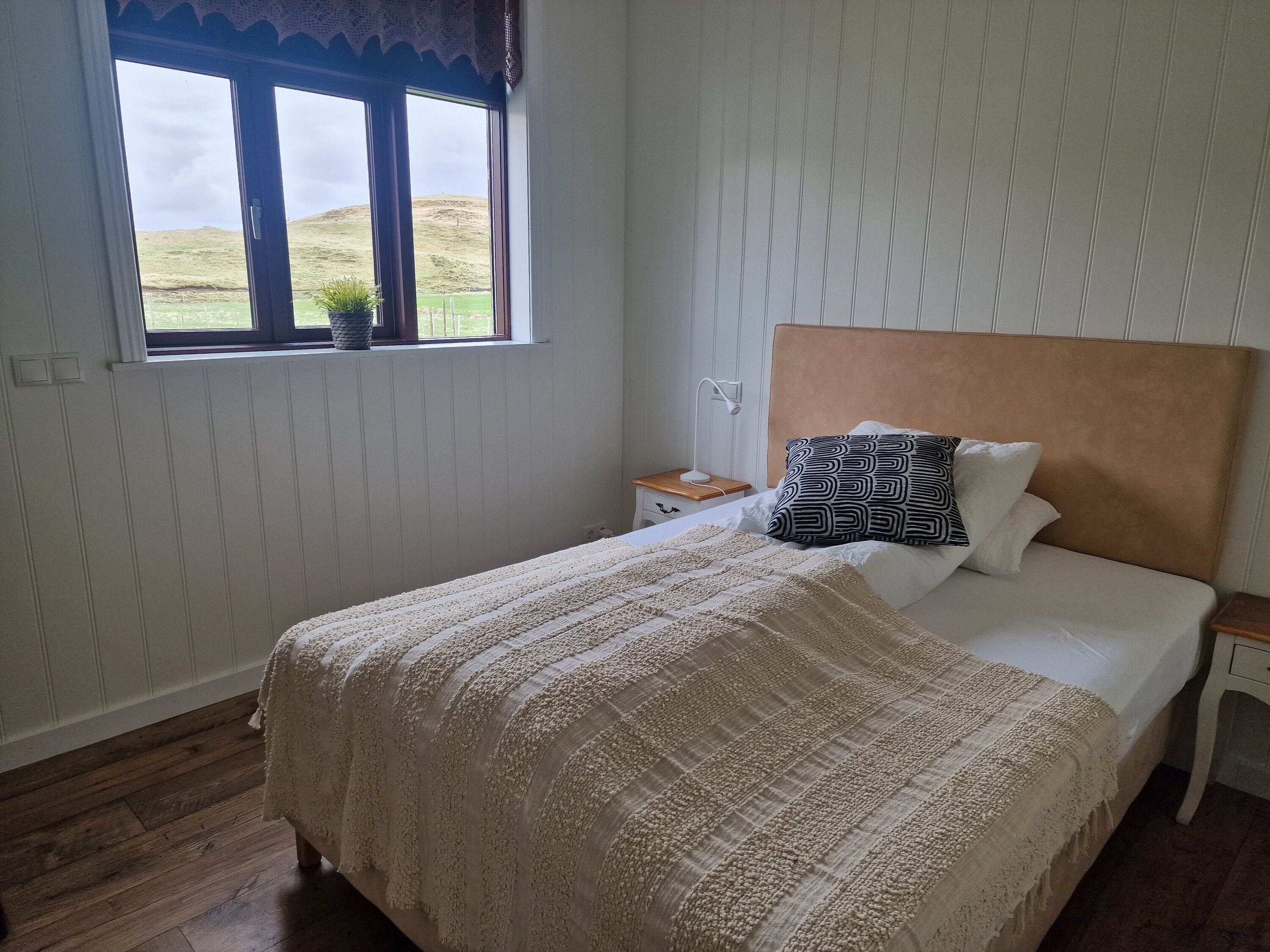 A bedroom with a large bed, a window with a view of hills, a bedside table with a lamp, and a nightstand.