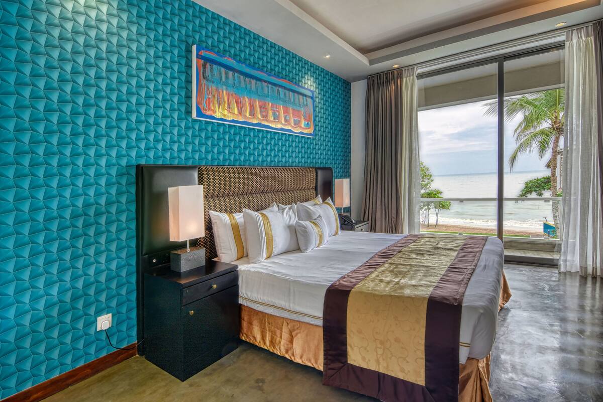 A hotel room with a large bed, a nightstand, a wall with a geometric pattern, and a view of the ocean through a large window.