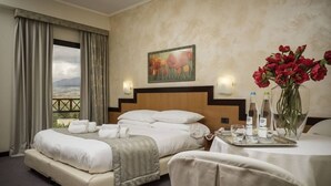 Quarto de hotel com cama, escrivaninha com um vaso de flores, um quadro na parede e vista pela janela.