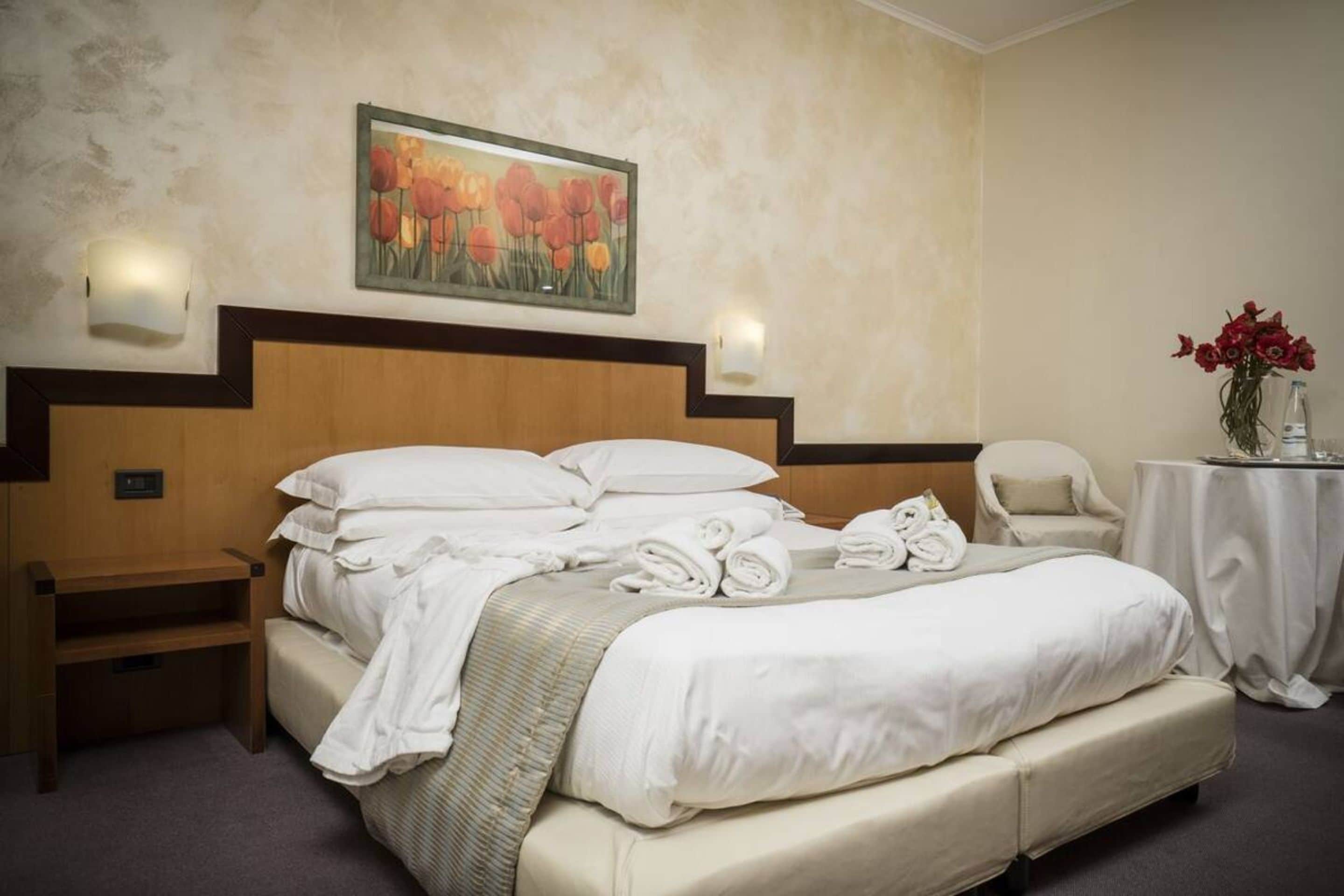 Une chambre d’hôtel avec un grand lit, une table de chevet, une chaise, une petite table ornée d’un vase de fleurs et un tableau accroché au mur.