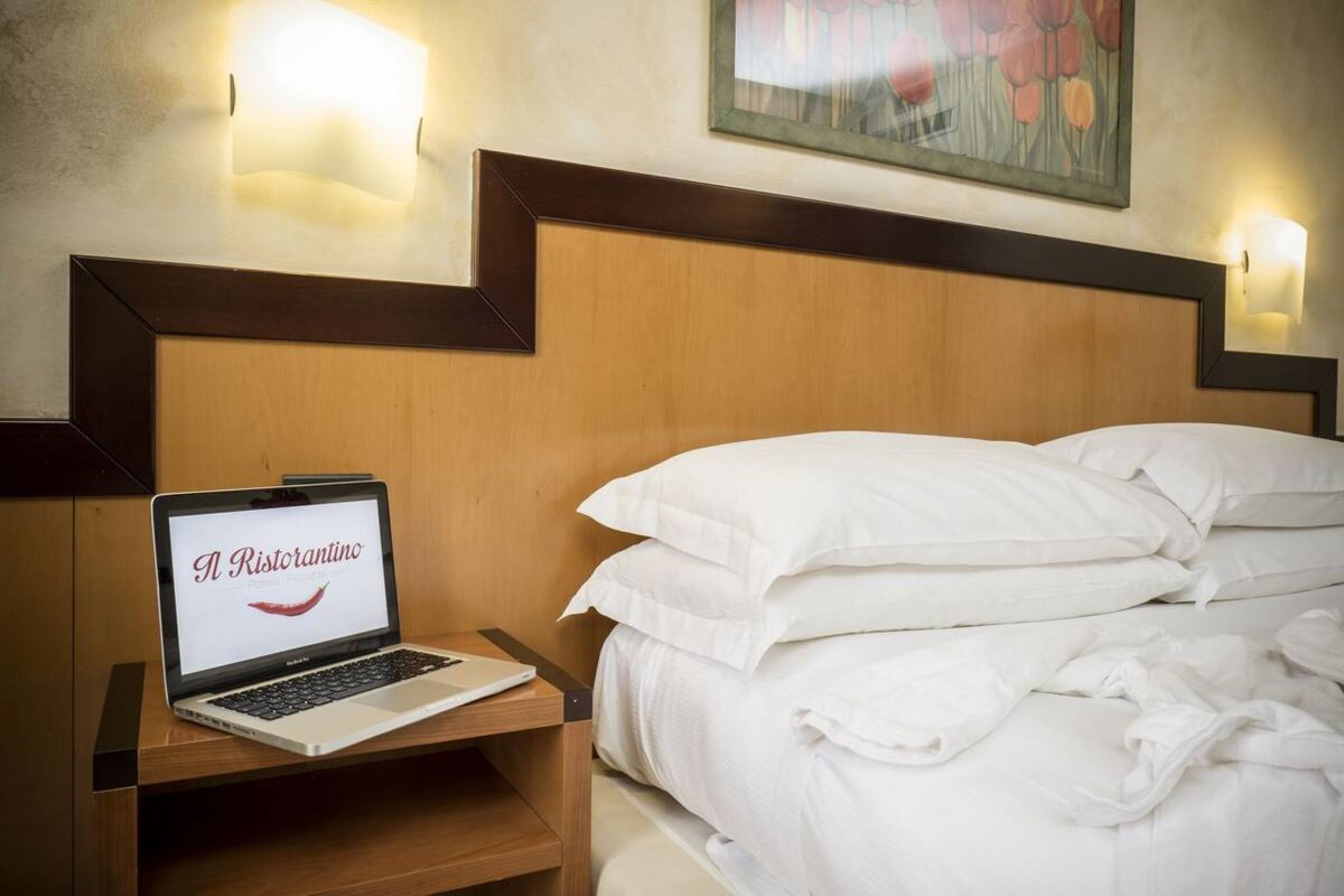 Une chambre d’hôtel avec une tête de lit en bois, un ordinateur portable sur une table de chevet et un lit recouvert de draps blancs.