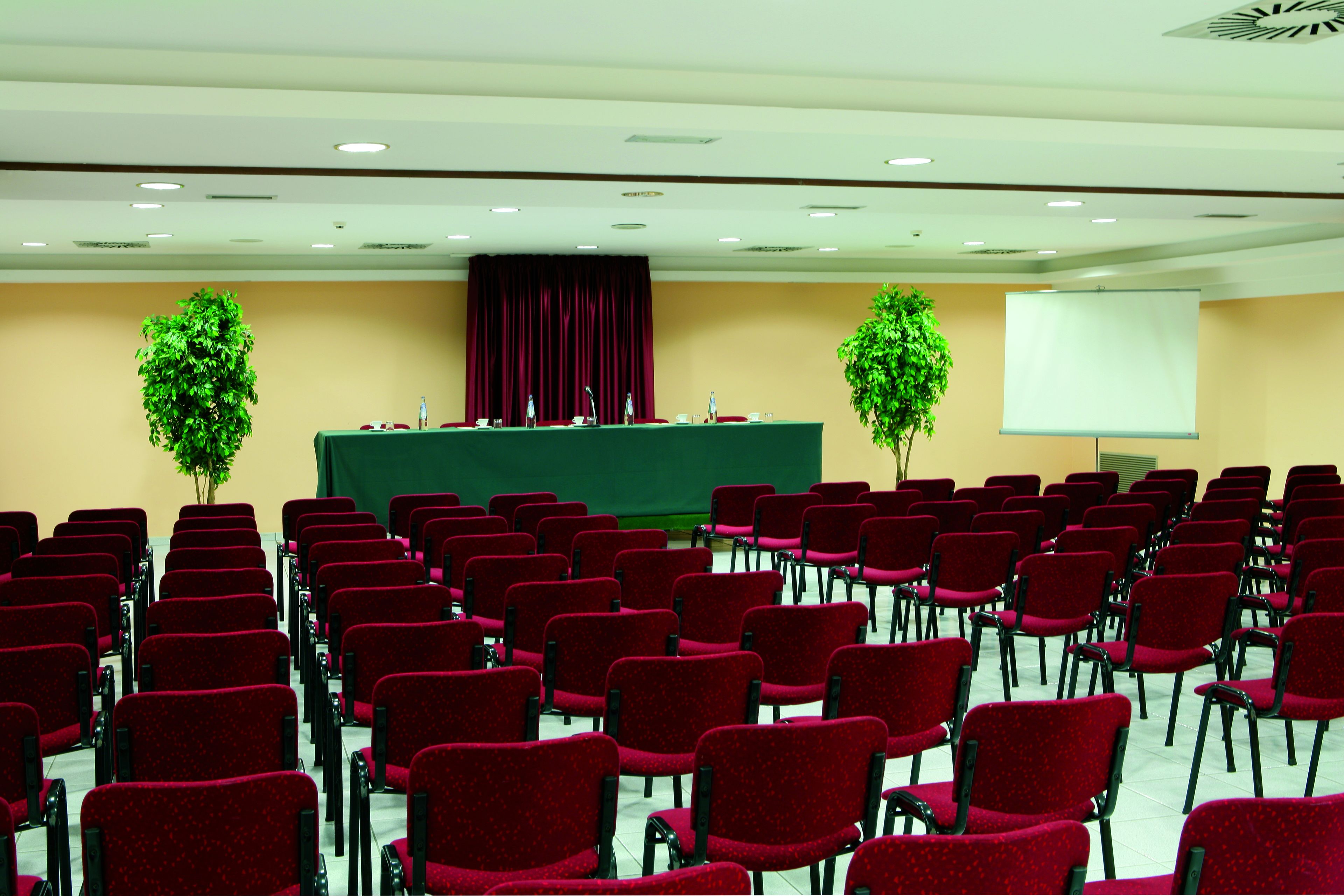 A conference room with rows of red chairs, a green table with a microphone, two potted plants, and a projection screen.