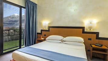 A hotel room with a large bed, two bedside tables, a nightstand, a window with curtains, and a view of a mountain.