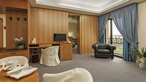 Quarto de hotel com escrivaninha de madeira, televisão, poltrona de couro e área de jantar com mesa posta para dois.