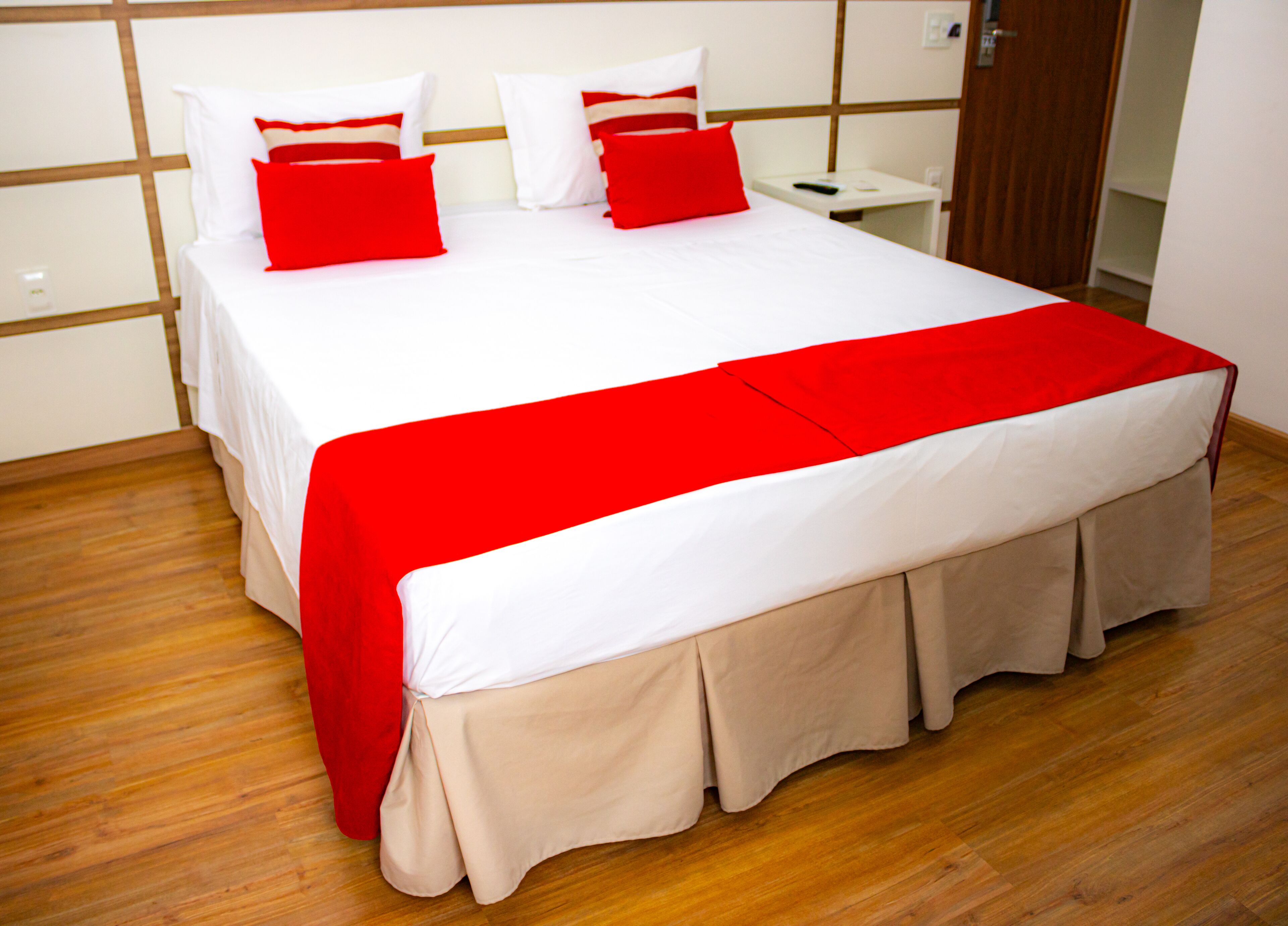 A neatly made bed with a red and white bedspread, a wooden floor, and a white wall with a wooden panel design.