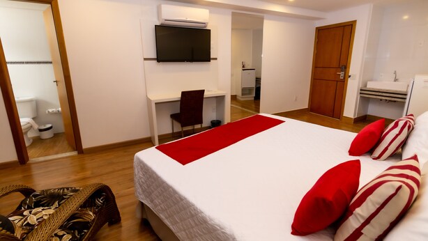 Quarto de hotel com cama, escrivaninha, cadeira, televisão, guarda-roupa e banheiro.