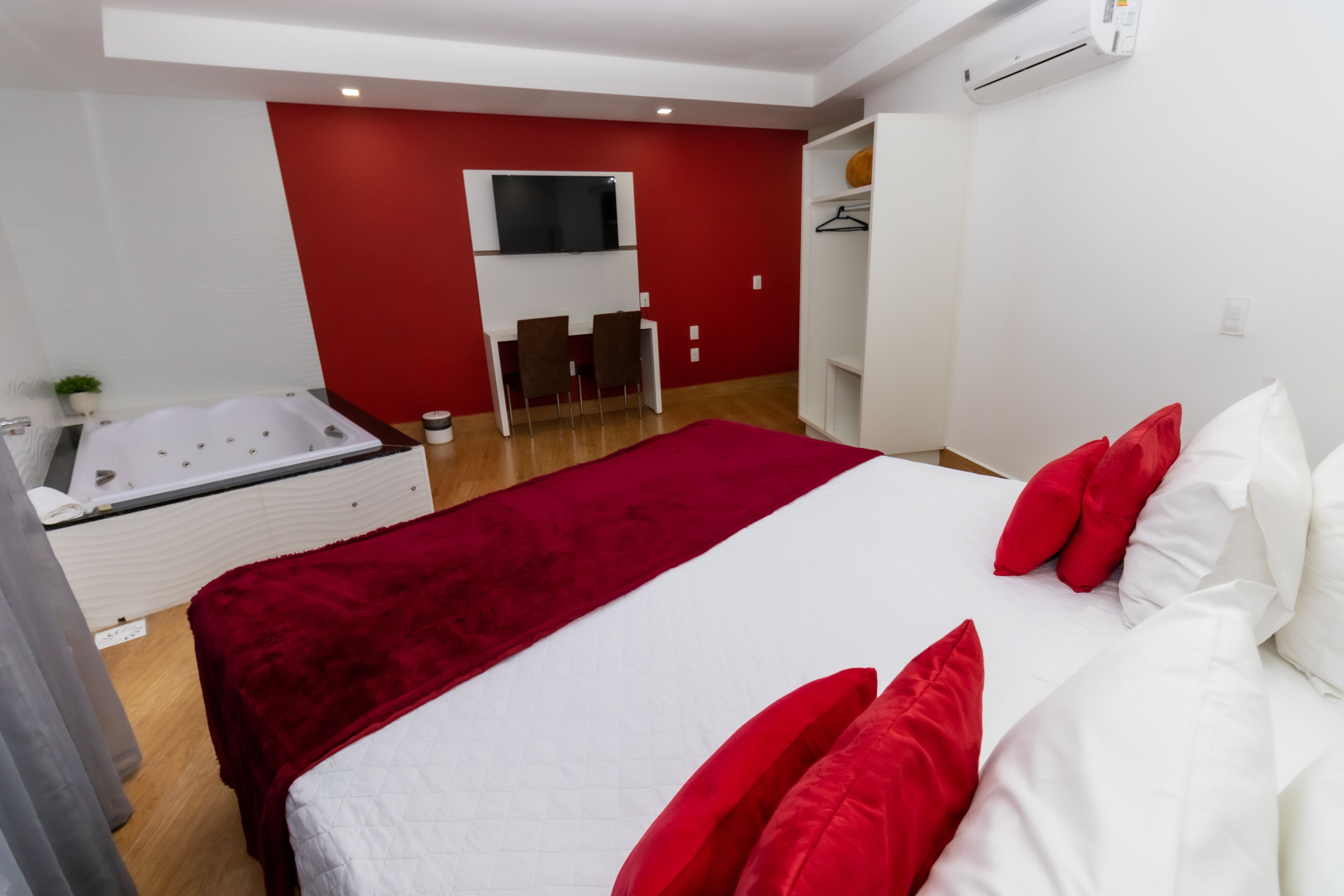 A modern bedroom with a large bed, a red accent wall, a TV mounted above a seating area, and a white wardrobe.