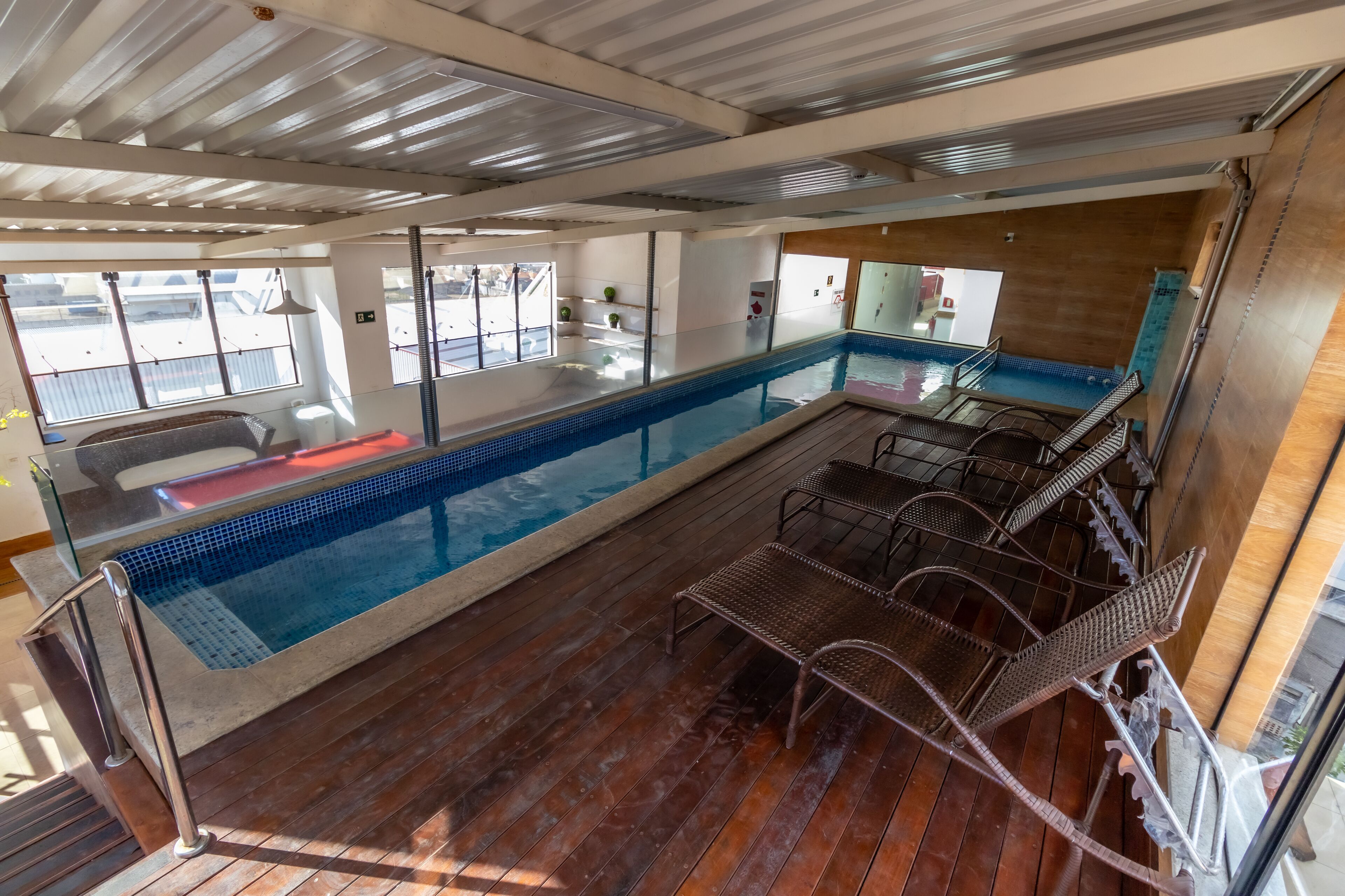 Zona de piscina con cubierta de madera, sillas de metal y una barandilla de vidrio.