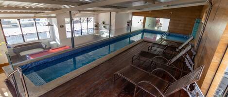 Zona de piscina con cubierta de madera, sillas de metal y una barandilla de vidrio.