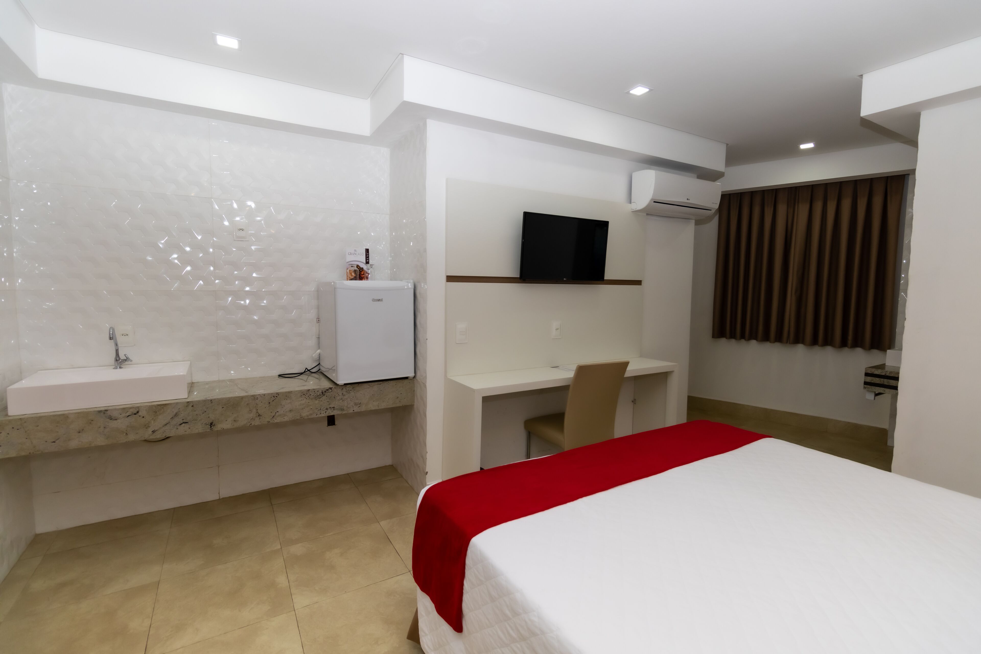 Quarto de hotel com cama, escrivaninha, cadeira, televisão, mini-frigorífico e banheiro com pia e espelho.