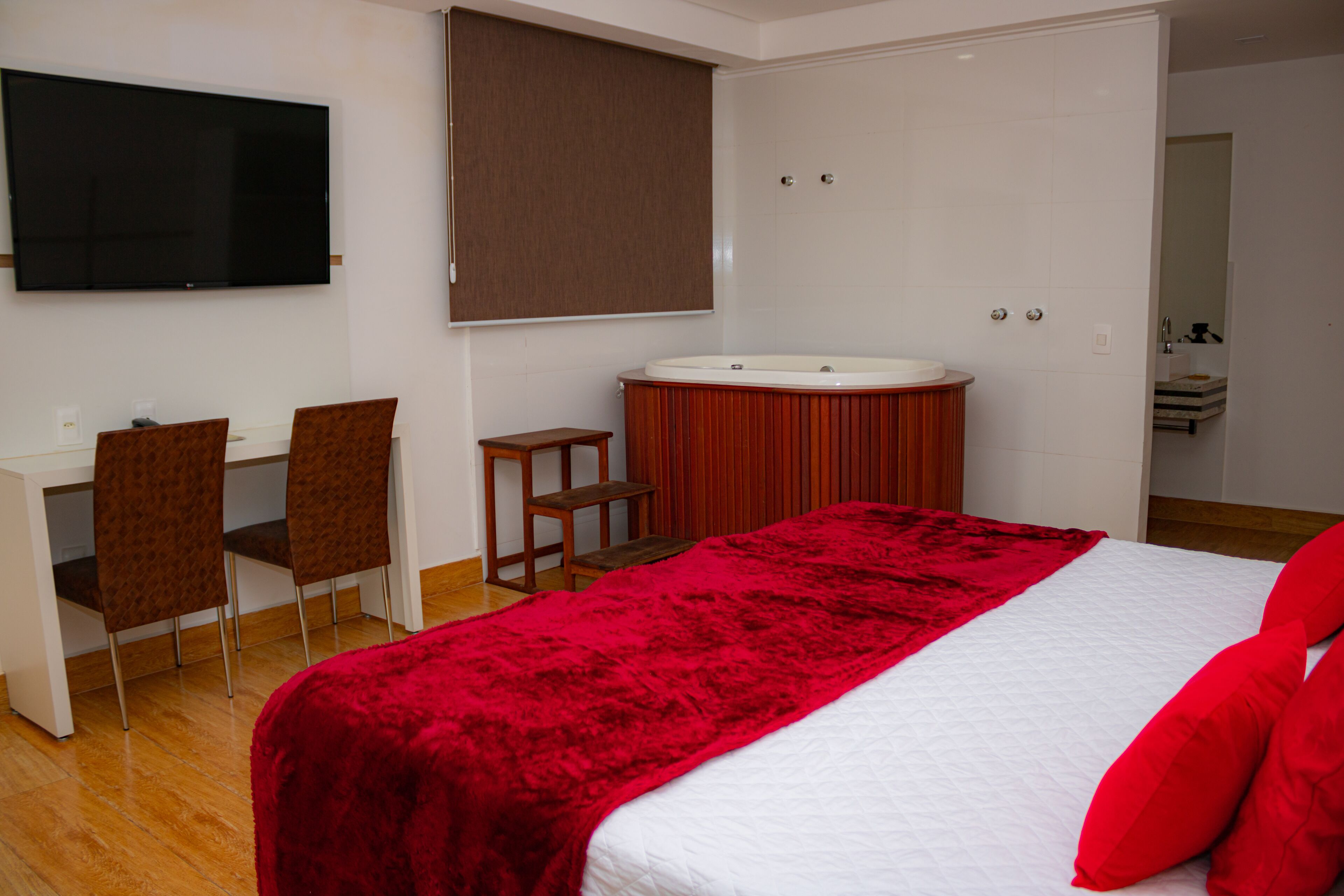 A bedroom with a bed, a red blanket, a TV, a desk with chairs, and a bathtub.