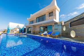 A modern poolside area with a clear blue pool, lounge chairs, and a two-story building with large windows. - Villa Paris Hill Villas Kalkan (Kas)