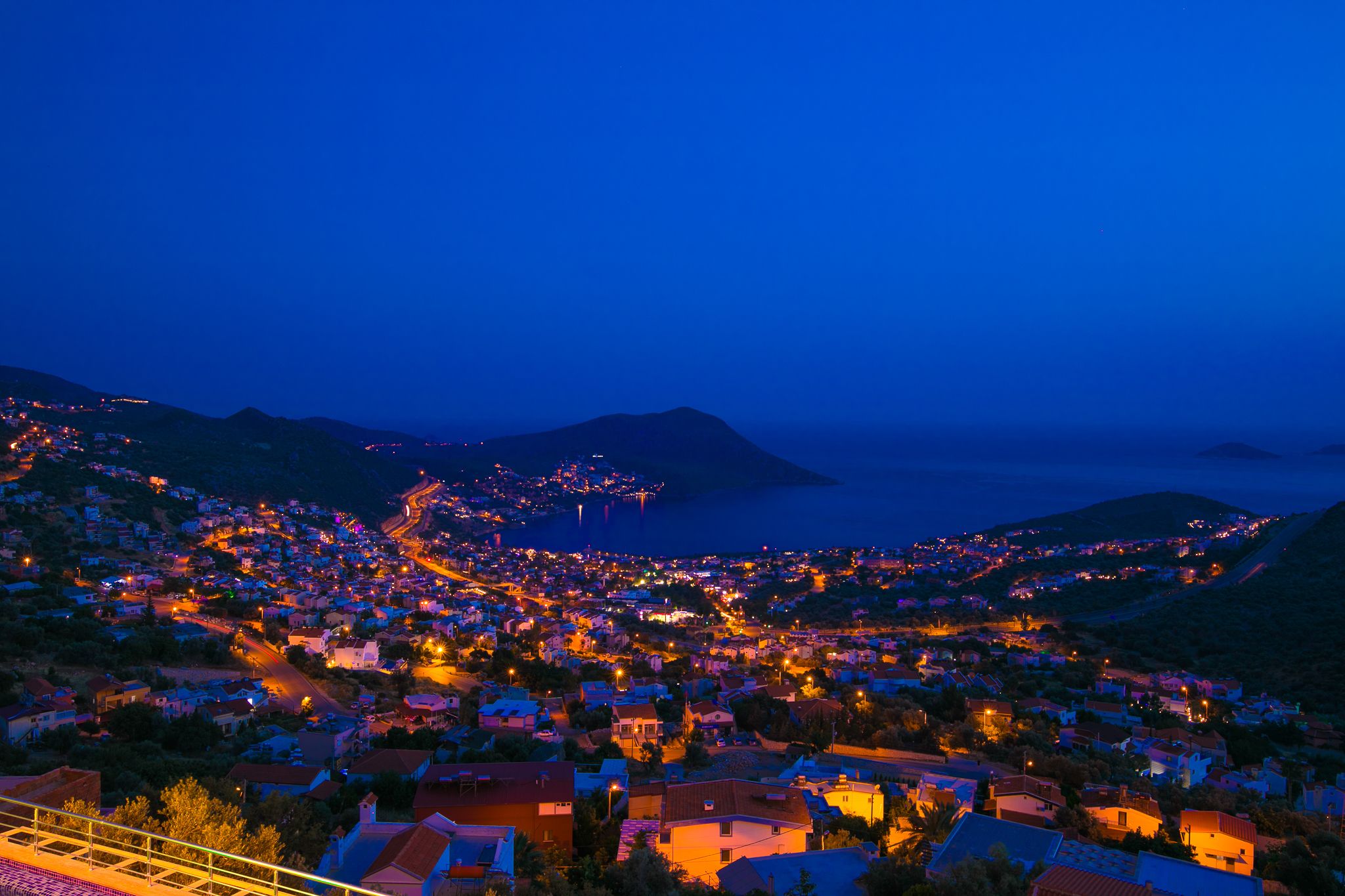 A coastal town at dusk with illuminated streets and buildings, a prominent hill in the background, and a view of the sea.