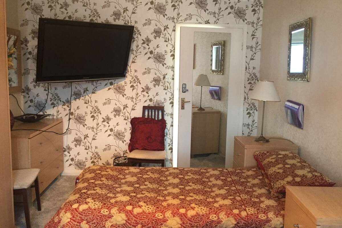 Un dormitorio con cama, escritorio, silla, televisor, espejo y una puerta que da a otra habitación.