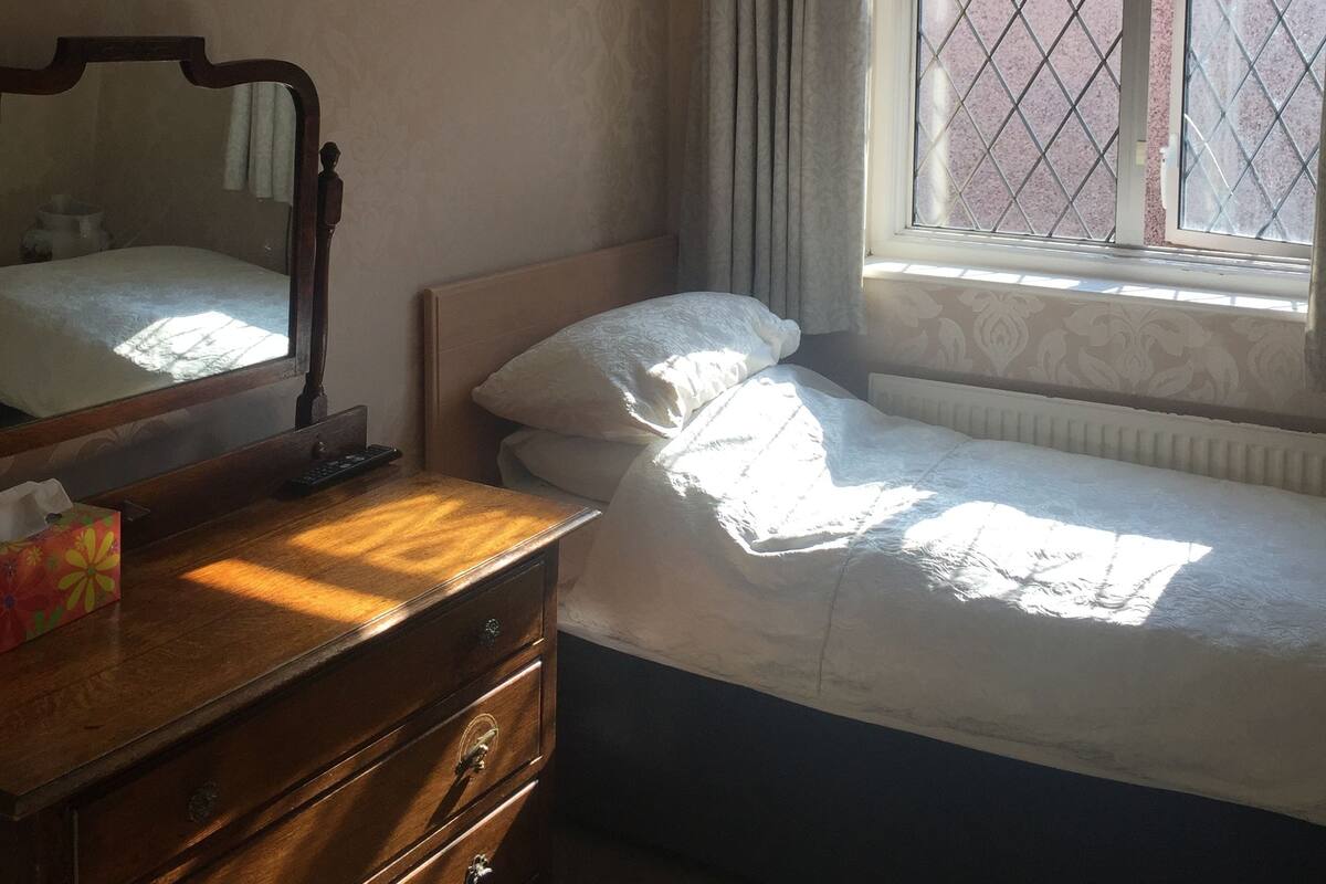 Un dormitorio con una cama individual, un aparador de madera, un espejo, una ventana con cortinas y una lámpara colgante.