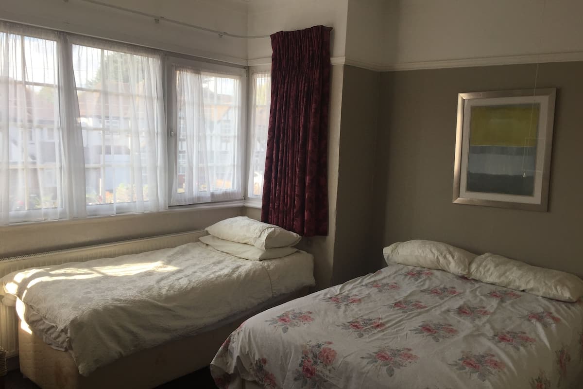Una habitación con dos camas, una con colcha estampada y la otra con sábanas blancas lisas. Se ve una ventana con cortinas translúcidas y una cortina rojo oscuro medio bajada. Un cuadro enmarco cuelga en la pared.