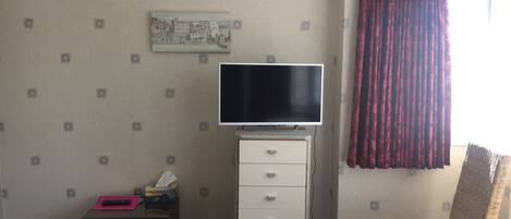 Una habitación con una televisión sobre un mueble, una mesa pequeña con objetos, una silla y una ventana con cortinas.