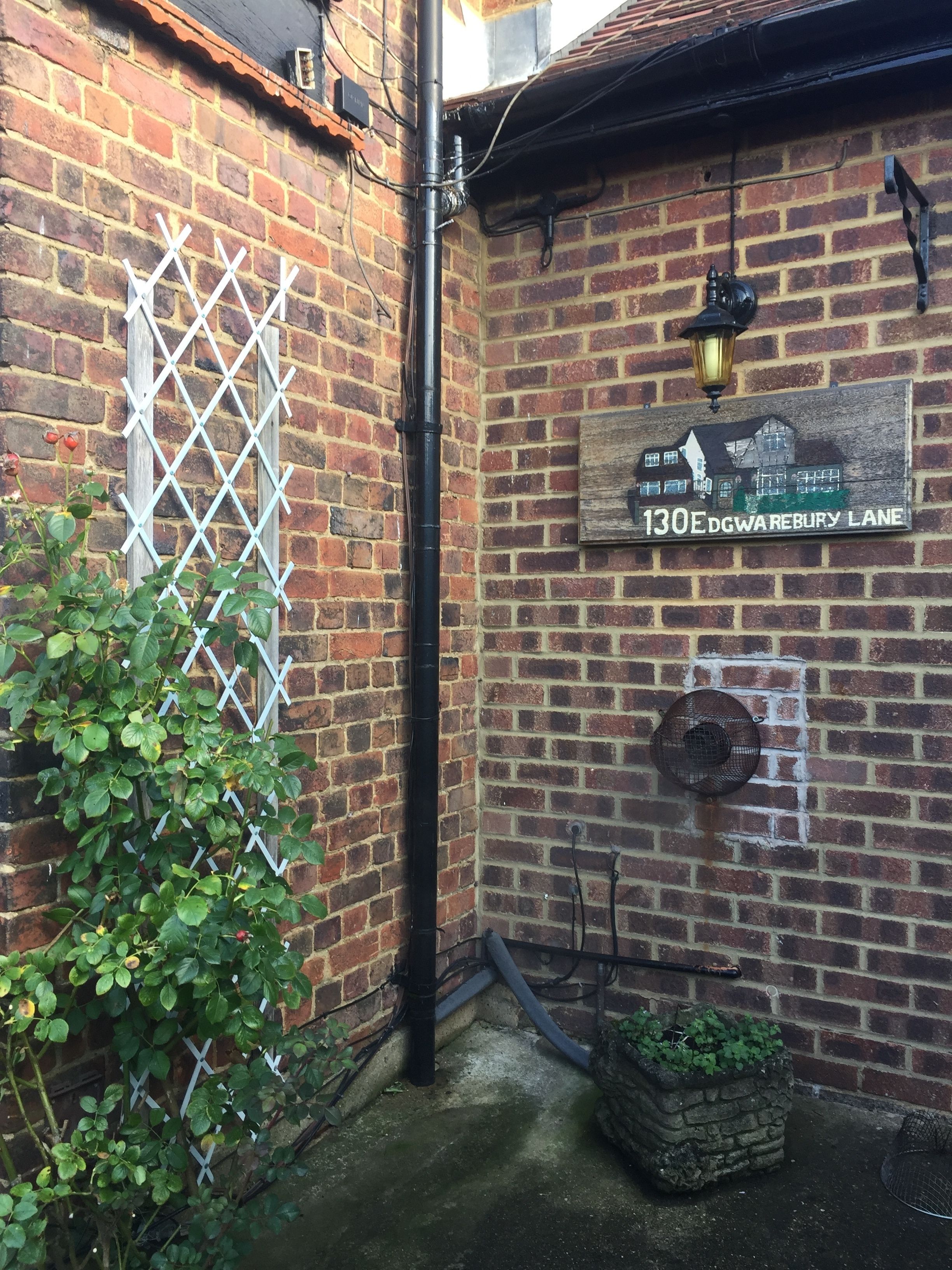 A brick wall with a white lattice window, a black pipe, a street sign reading "130E DGWAREBURY LANE", and a small garden area with green plants.