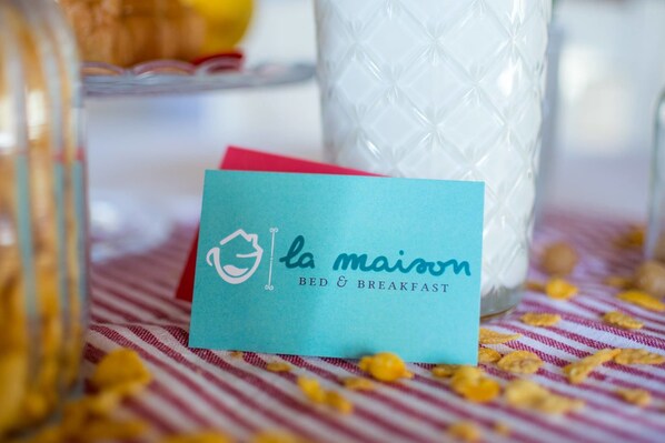 A card with the text "la maison BED & BREAKFAST" on a table with a striped cloth and scattered cereal.