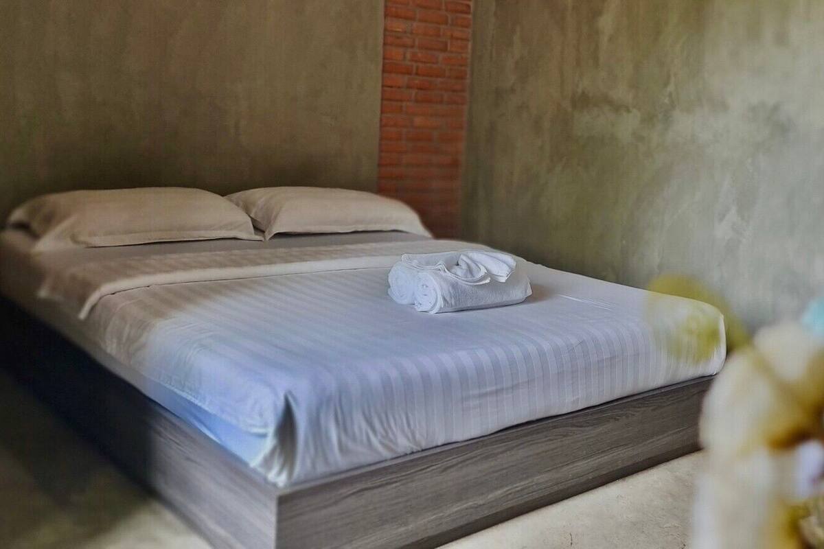 Un lit simple avec un cadre en bois et une couvre-lit grise, un mur de briques derrière, et une plante au premier plan.
