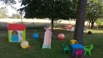 Un espacio al aire libre con césped, una casita de juegos, un tobogán, sillas de plástico de colores y una hamaca.