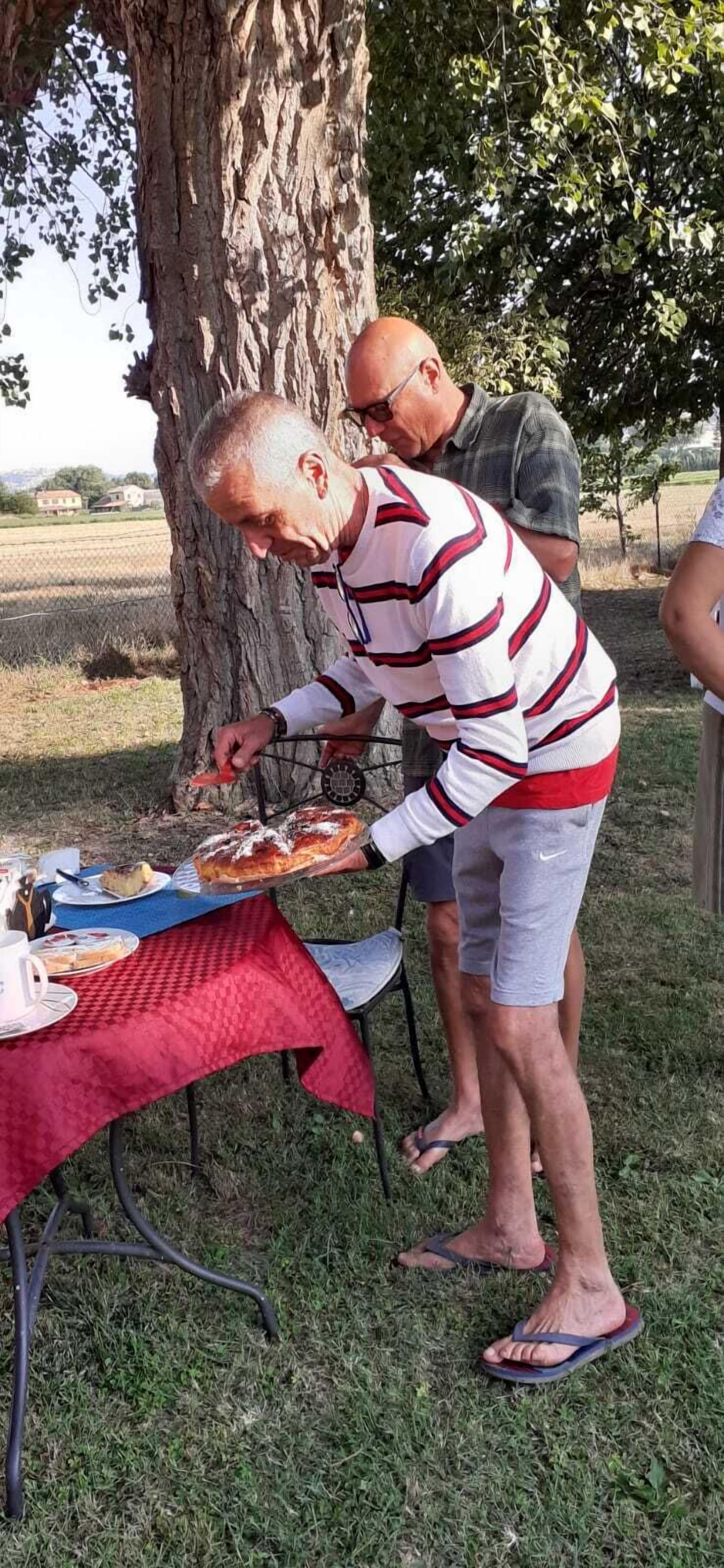 Un homme sert des plats à une table en plein air, recouverte d’une nappe rouge, et l’ensemble est entouré d’arbres.