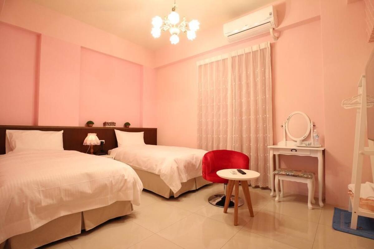 A hotel room with two beds, a red chair, a small table, a vanity with a mirror, and an air conditioning unit.