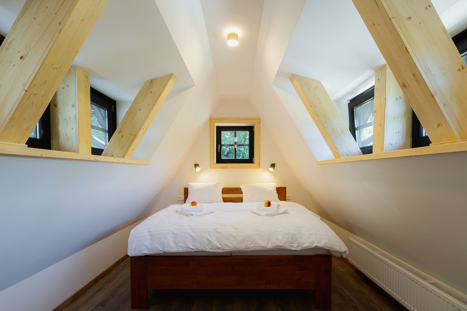 Une chambre avec un plafond en pente, des poutres en bois, un lit avec du linge de maison blanc, une petite fenêtre et une lampe fixée au mur.