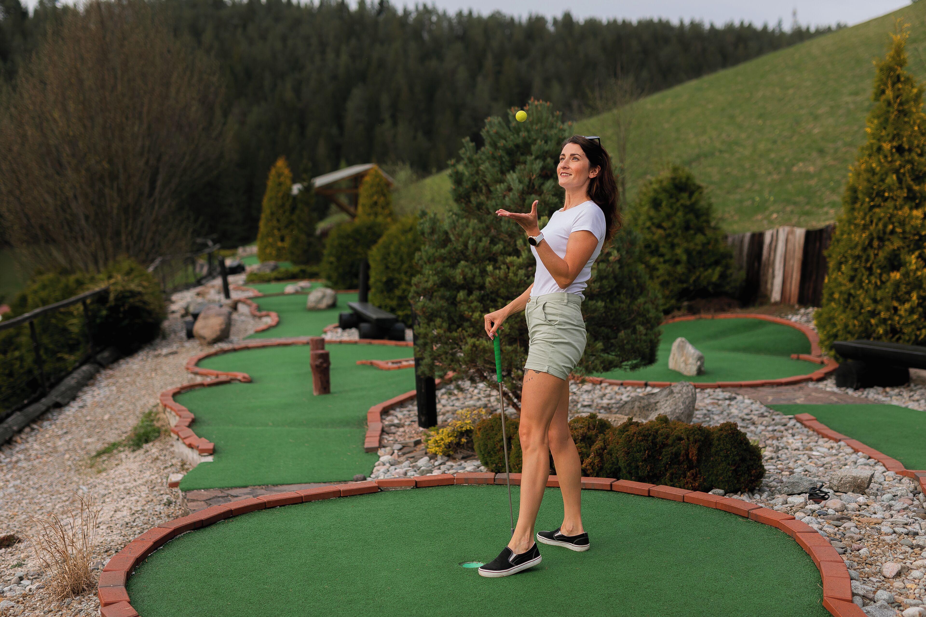 Iemand speelt minigolf op een groen veld, met een houten bankje en bomen op de achtergrond.