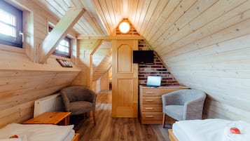 Una camera da letto mansardata accogliente, con pareti in legno, soffitto a falde inclinate, un letto, due poltrone, una scrivania con un laptop e una TV a parete.
